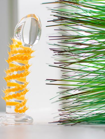 The Taffy Tickler Silicone Sweets glass dildo, covered in little orange spikes, standing upright near some pointy green fake grass.