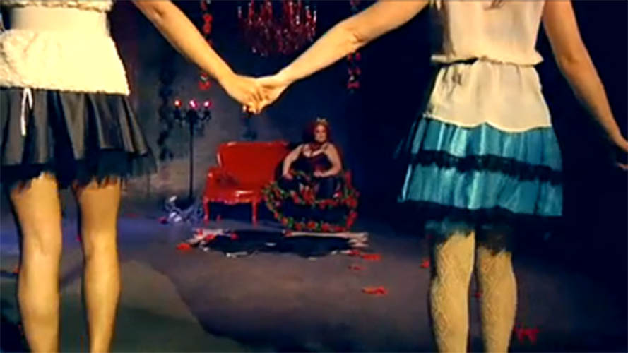 Two women holding hands in front of the Queen of Hearts, April Flores, sitting on her throne.