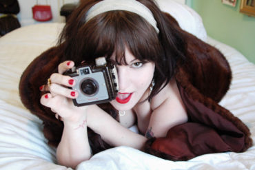 Courtney Trouble on a bed, holding a camera up to their eye.