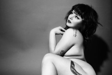 Courtney Trouble, nude, in a black and white shot showing off their high heel tattoo.