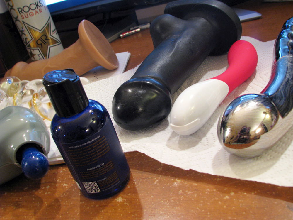 My desk, covered in messy used dildos and vibrators, with a bottle of lube in the foreground.