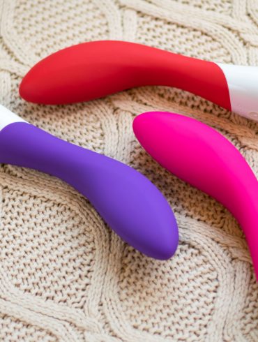 The LELO Mona 2 rechargeable vibrator in three colors: purple, red, and cerise (pink), lying on a cream-colored knit fabric.