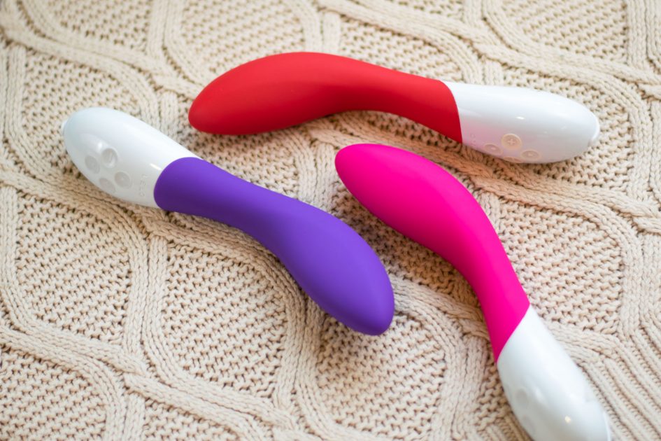 The LELO Mona 2 rechargeable vibrator in three colors: purple, red, and cerise (pink), lying on a cream-colored knit fabric.