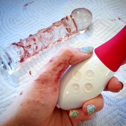 My menstrual bloody hand holding the LELO Mona 2, with glass dildo in background.