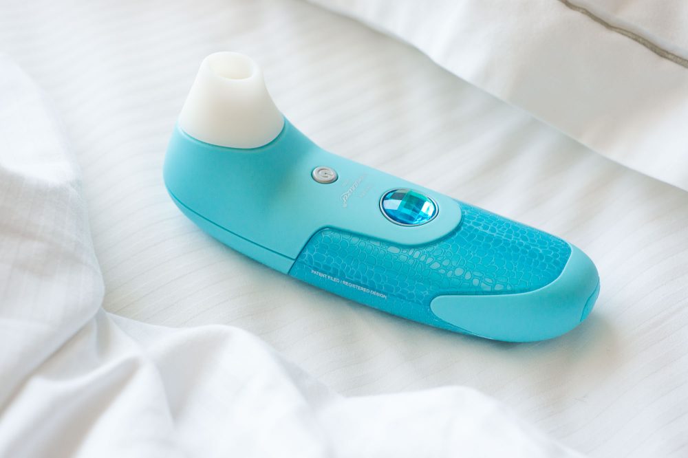 Womanizer air flow / pressure wave suction vibrator, with its ridiculous fake jewel, on a very white bed.