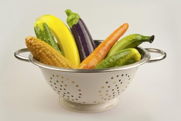 A literal colander full of silicone sex toys shaped like various vegetables and fruit: corn, carrot, eggplant, banana...