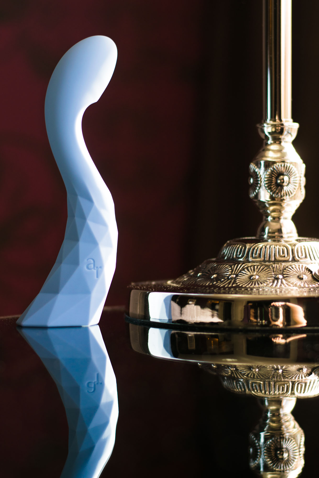 L'Amourose Prism V rechargeable G-spot vibrator standing up on a reflective table, next to an ornate golden lamp.