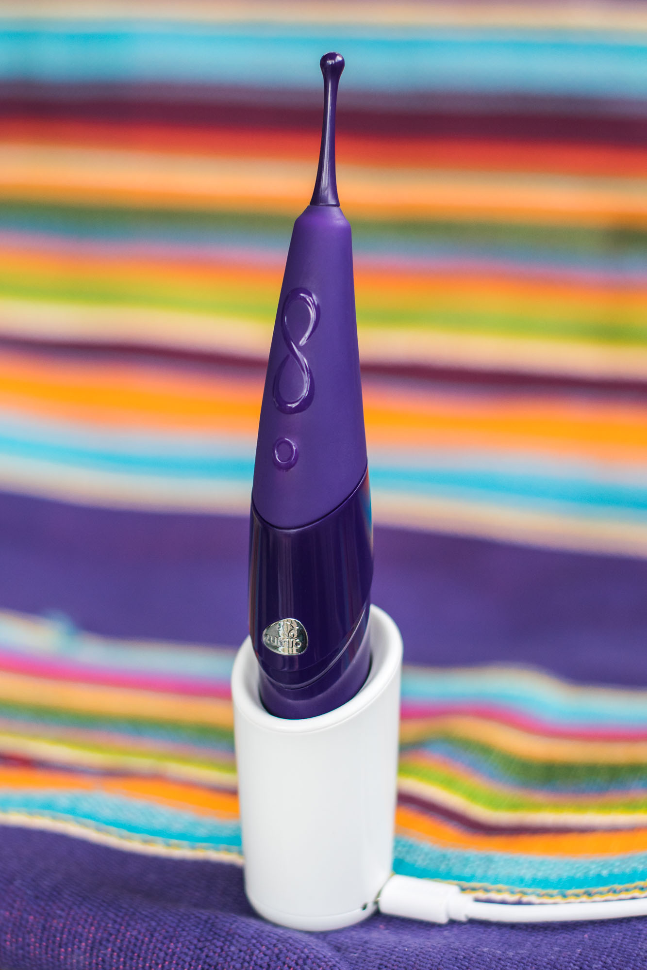 Zumio X rechargeable clitoral vibrator, in its charging dock, on a colorful striped blanket.