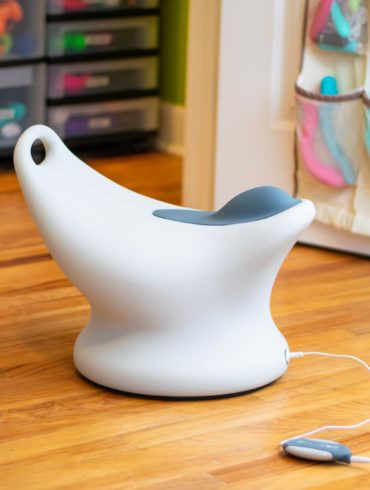 The Jimmyjane Rumble Seat, a white plastic toilet-looking chair with a grey vibrating bump on top, standing in front of my sex toy closet.