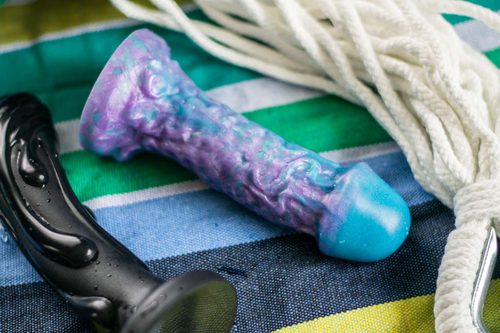 Tantus Magma (left) and Uberrime Jellyfish 2.0 silicone dildos lying on colorful striped canvas, next to some rope.