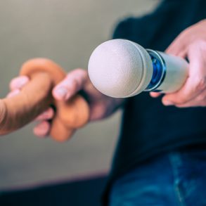 A headless man holding a Magic Wand and a realistic dildo toward the camera.