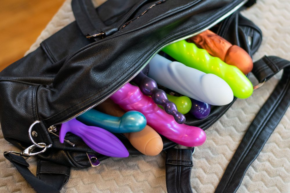 A black leather bag with a bunch of colorful silicone dildos spilling out.