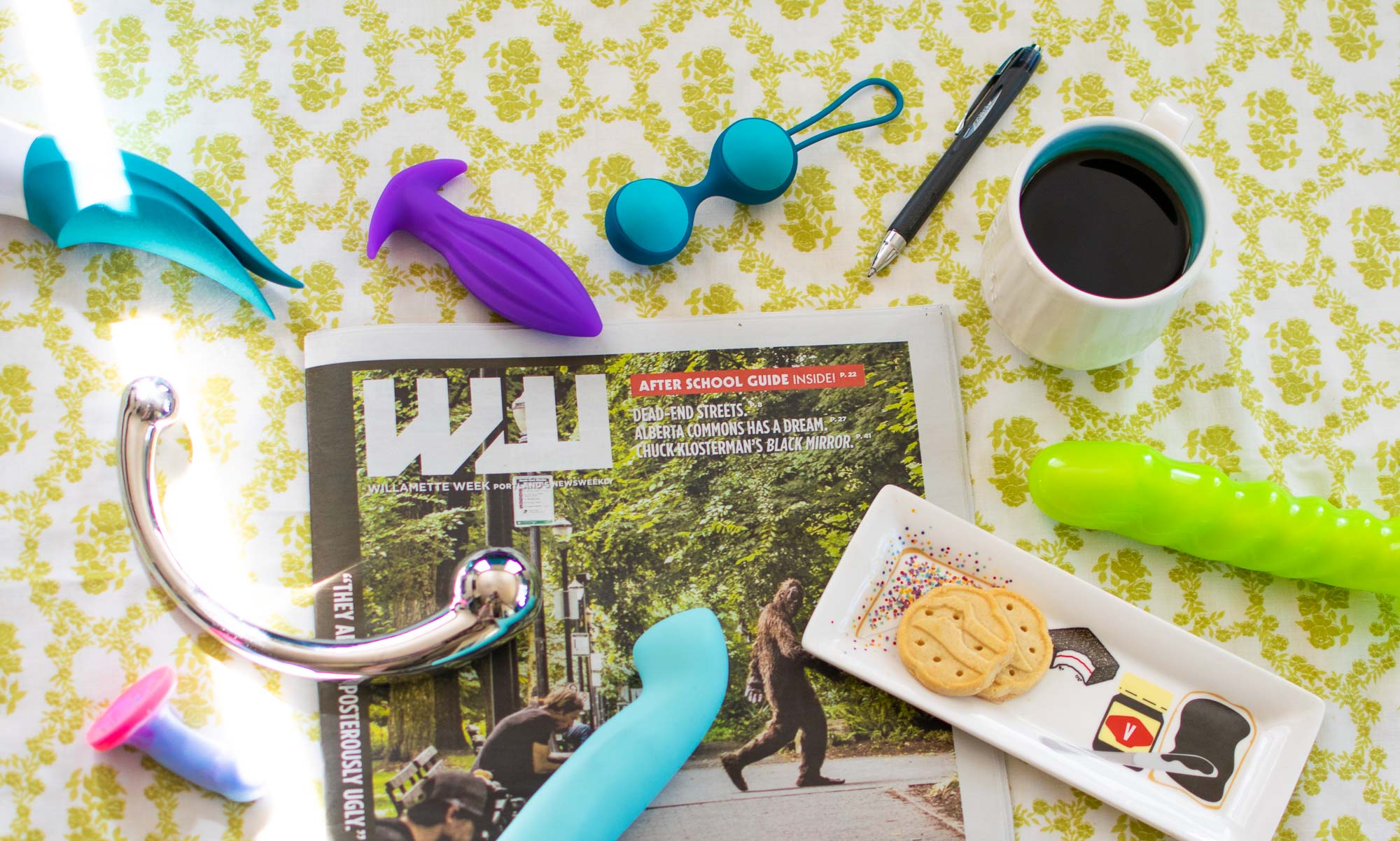 From-above shot of a newspaper, with a cup of coffee, a platter with cookies, and sex toys all around.