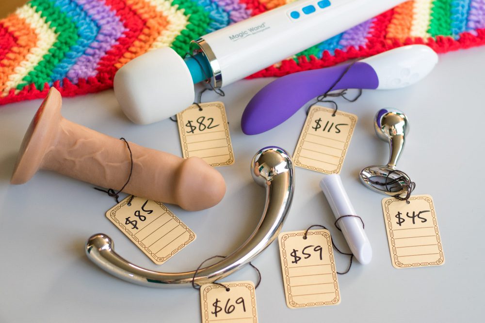 Various sex toys laid out on a grey table next to a rainbow blanket, with hang tags displaying handwritten prices.