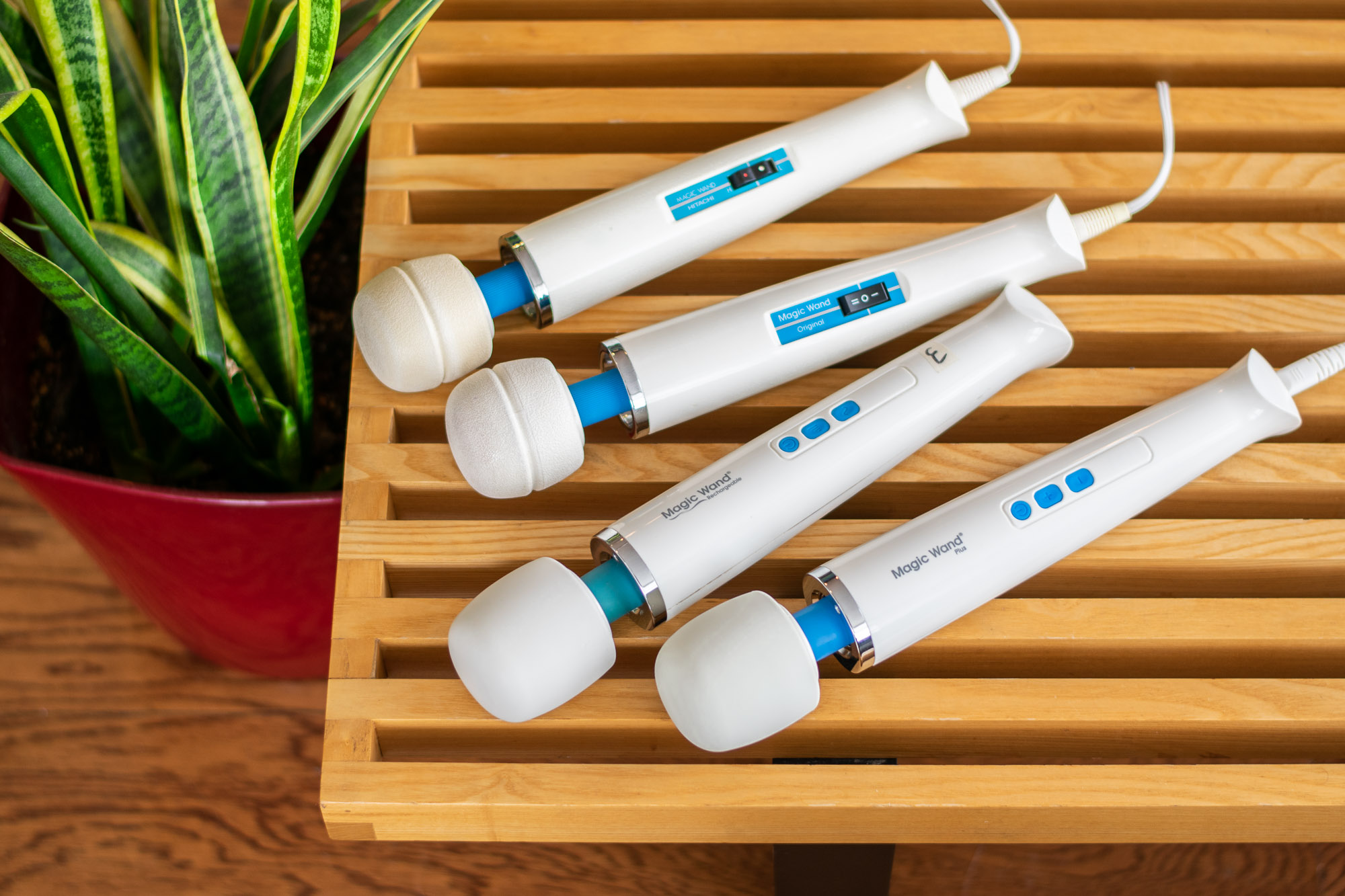 Four different iterations of the Hitachi Magic Wand vibrator, lying on top of a wooden bench near a green plant.