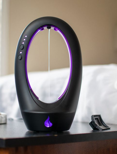 Warm Touch automatic warming lube dispenser standing upright and continuously dispensing, next to a lamp that resembles anal beads, and my cat on the bed.