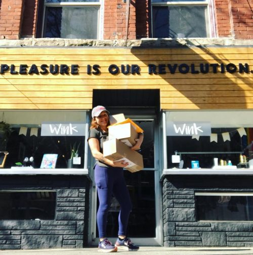 Jenn Mason, owner of WinkWink, standing outside the shop holding a bunch of packages. The shop behind her reads, "PLEASURE IS OUR REVOLUTION."