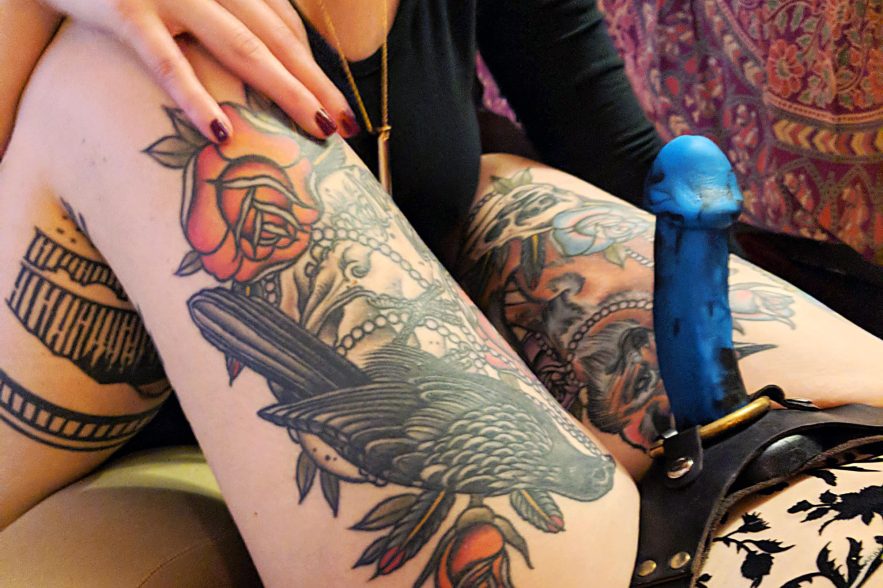 A hand caressing the tattooed leg of a person wearing a strap-on: the New York Collective Shilo dildo.