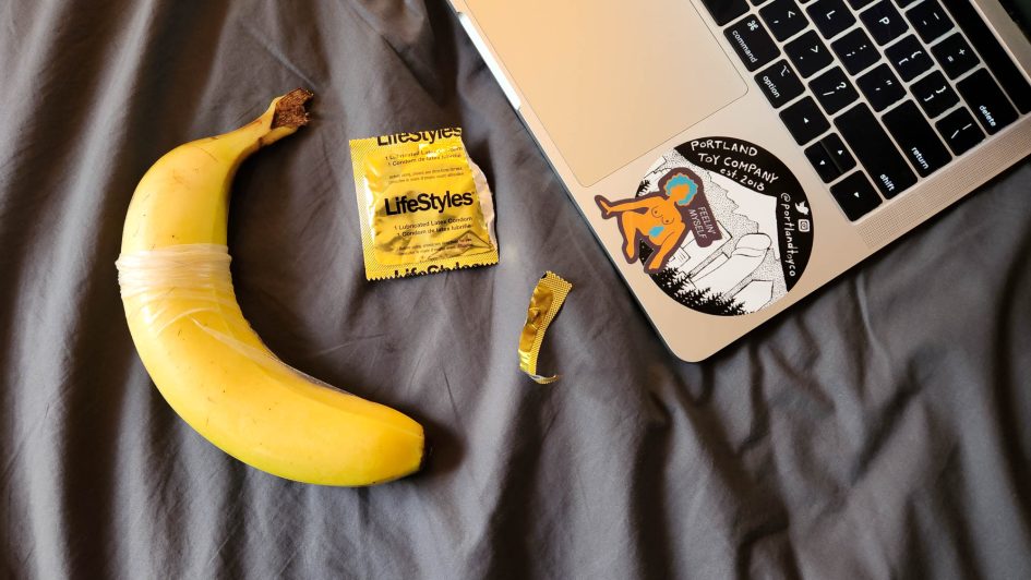 On top of my sheets, a banana with a condom on it next to my laptop and an open condom wrapper.
