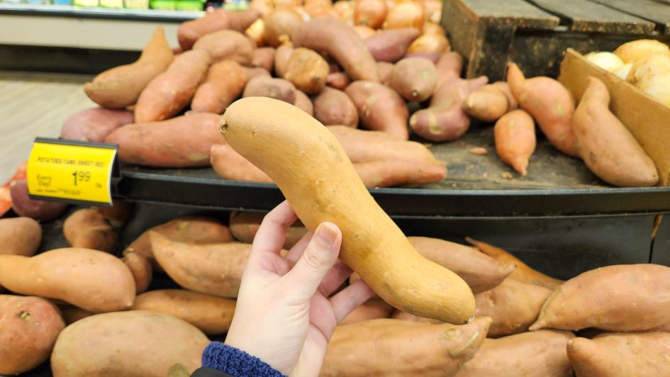 Me holding my chosen yam, which is shaped like an extremely lazy S, in front of piles of other yams at the grocery store.
