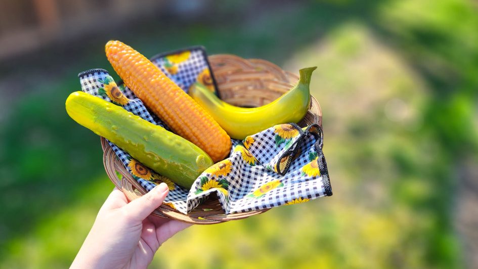 Me holding a basket containing three silicone dildos shaped like a cucumber, corn on the cob, and banana.